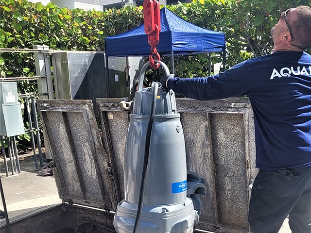 Man looking up helps to guide placement of lift station pump hanging from red hook into an opening with metal hinged doors lifted