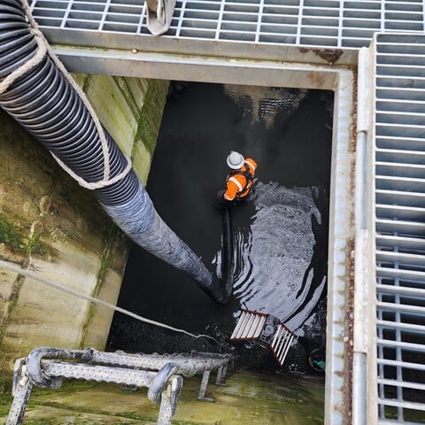 View down a street-level opening, past the grate, wall-mounted metal ladder and flexible pipe to construction worker standing in water at the bottom